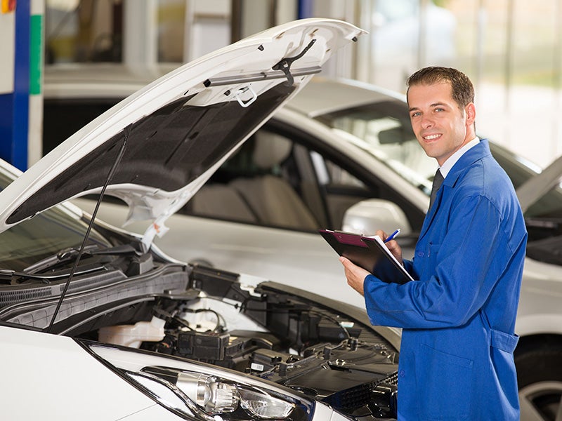 A service advisor in a blue coat fills out a clipboard by an open car hood.